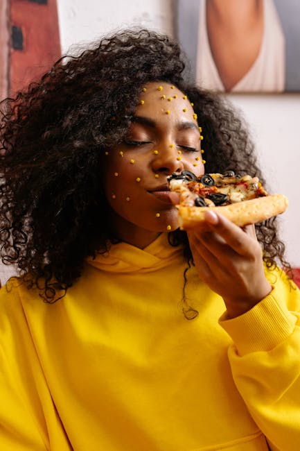 Woman wearing a yellow hoodie enjoying a slice of pizza with creative face art indoors.