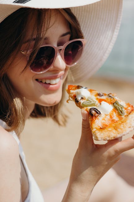 A woman in a hat and sunglasses enjoys a slice of pizza on a sunny day.