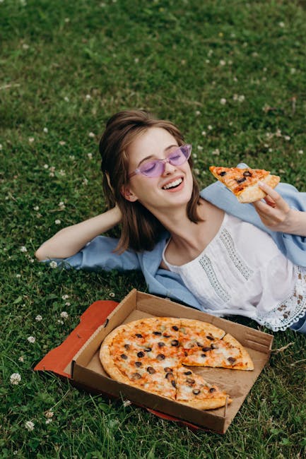 Young woman relaxing on grass, savoring pizza from a box while smiling softly.